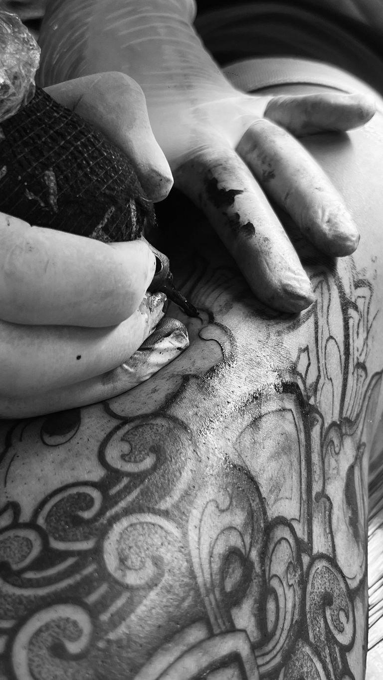 Black and white image of a tattoo artist working on a detailed design on a client's leg.